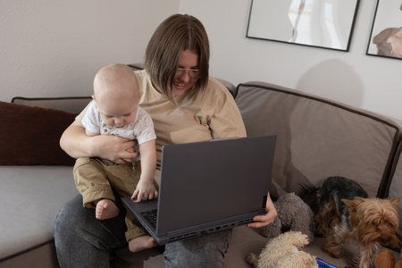 mother holding baby at laptop on sofa, smiling while working on call with dog nearby and cozy livingroomの写真素材