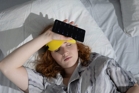 white woman reclining with remote and eye mask, remote held near face, relaxed posture on bed, streaming and screen culture vibe, cozy slow content consumption and leisure modeの写真素材