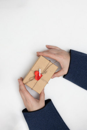 Two hands hold a small gift box tied with twine and a red heart decoration. The setting is simple and suggests a moment of giving for Valentines Day.の写真素材