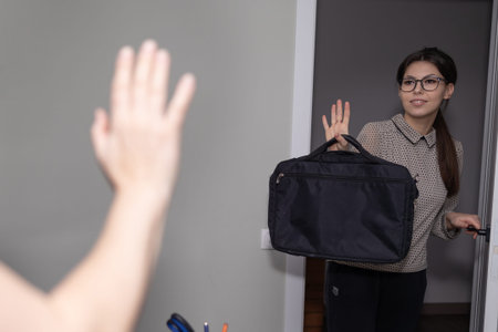 A young woman wearing glasses and holding a black briefcase is waving goodbye to someone while...