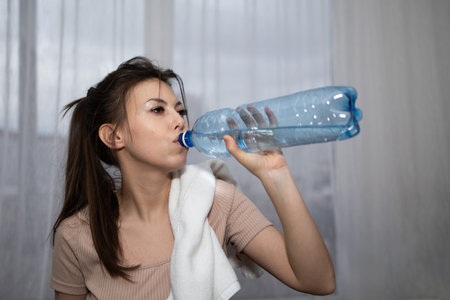 A young woman takes a drink from a large bottle of water. She holds a towel around her neck in a...