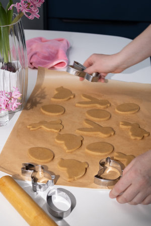 Hands cut out raw gingerbread dough into Easter bunny, egg, and chick shapes on baking paper. A vase...