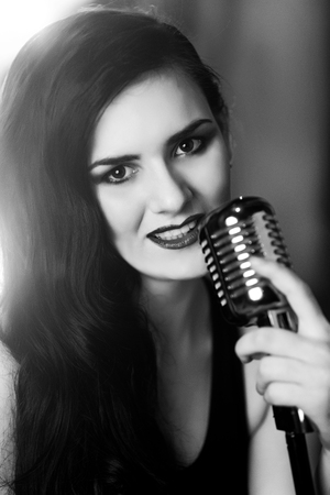 Beautiful woman with long black hair, deep breast cut jacket singing and holding big silver microphone, gray fog on background free place. Close up portrait. Sexy smile teeth smiling. Vertical bannerの写真素材