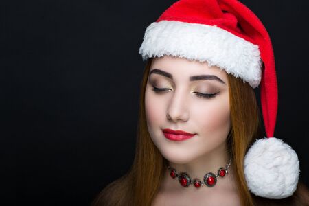 Young face closeup photo portrait. Beautiful girl wearing Christmas red hat cap. Pretty woman closed eyes stylish make up, happy New Year party. Santa girl, dreaming think about presents and surprisesの写真素材