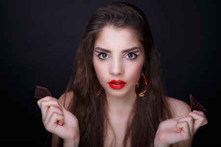 Beautiful woman, holding in her hands chocolate piece bar. Long hair brunette. Professional cosmetics makeup. Red matte lipstick lip-gloss. New photo close portrait, black color background horizontalの写真素材