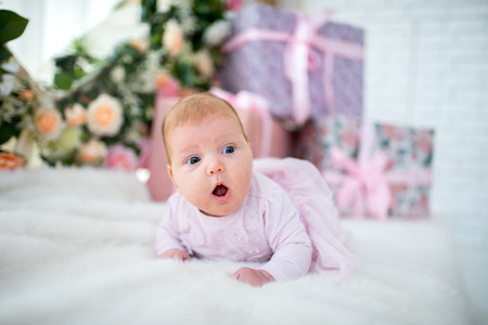 Beautiful baby girl crawling and rolling over in bed, white cover carpet. emotional pretty kid in pink dress. lie on stomach. horizontal background flowers, box with presents. family love concept の写真素材