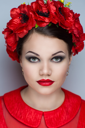Beautiful woman wearing big blossom wreath of red poppies. Spring, summer seasonal flowers. Closeup portrait of young girl woman styling Luxury Bright makeup shiny lipstick cosmetics. person accessoryの写真素材
