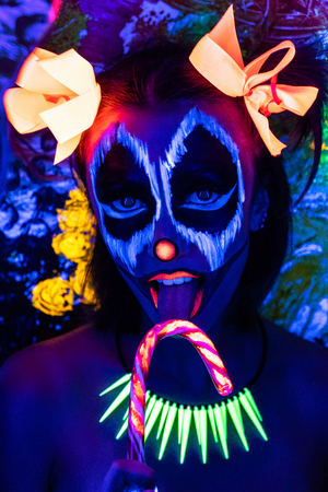 Scary clown make-up for Halloween. Cut skin on cheeks red nose, blood mouth flowing from wounds. Lollipop licking. Horrors terrible nightmares. Large orange bows hair tied in two ponytails. Funny faceの写真素材