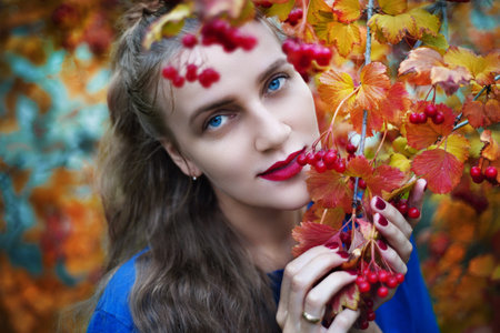 Young beautiful girl touch fruits of red viburnum in her hands. Autumn photo shows cozy mood, relax, tranquillity. Seasonal nature, picnic. Bright yellow leaves. Shiny blue eyes, matte red lipstickの写真素材