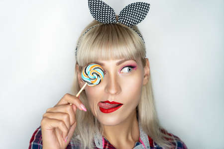 Beautiful makeup for a glamorous retro party. Pink shadows on white skin of the model, bright red lipstick. Large lollipop, round sucking candy is in the hand of the model, closes one eye. White hairの写真素材