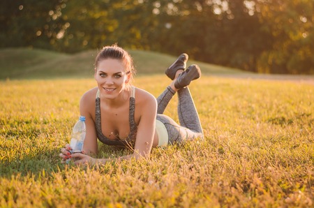 Attractive fitness woman in sportswear lies on the grass in the park and holding a bottle of water after exercise.の写真素材