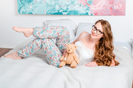 young pretty blonde girl in pajamas on the bed in his room and holds a soft toy in the hands and smilingの写真素材