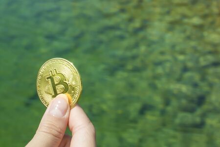 Bitcoin in a girl s hand against the background of a lake, grass, stones under water. Background with copy space for text. Prosperity, Growth, Financial Success.の写真素材