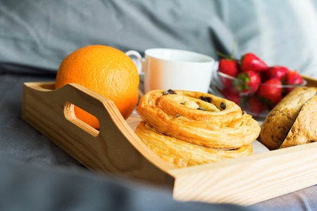 Morning breakfast in bed. Wooden tray coffee bun orange fruit coffee strawberry on grey linens bedding sheet pillow coverlet in gray colors hotel room interior. Copy spaceの写真素材