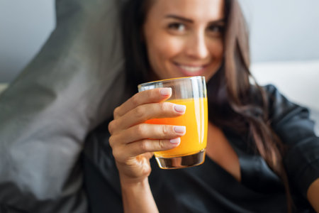Breakfast in bed. Young beautiful brunette woman in black silk dressing gown drink orange juice....
