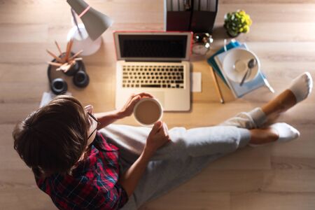 Woman is sitting on floor taking a break with coffee and working on computer remotely at home.の写真素材