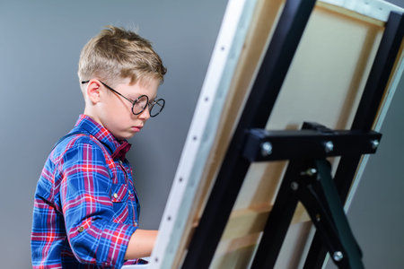 Portrait of cute schoolboy artist in eyeglasses painting picture on canvas