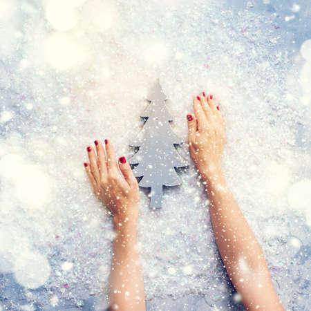 Card with Festive Vintage Christmas Wooden Fir Tree Female Hands Holding One Holiday Decorative Object on Snow Background Top View Flat Lay Advent Gift Toned Light Effectの写真素材
