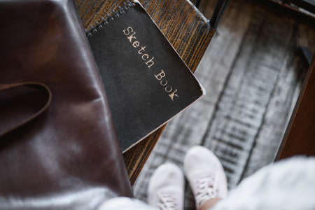Sketchbook, leather bag and feet in boots on wooden white floor. View from above. spend your free time qualityの写真素材