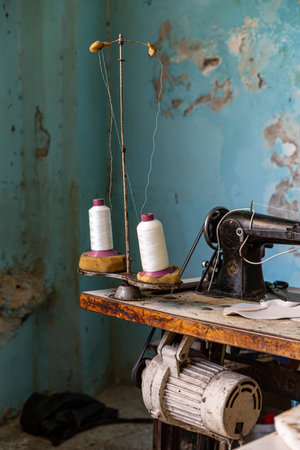 Old sewing machine on a worn wooden table with two white-thread spools and a weathered teal wall,...