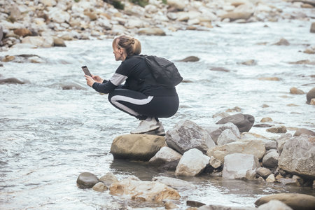 a blonde woman blogger in sports comfortable clothes and with a backpack on her shoulders takes pictures of nature in the mountains and a mountain river with a course on her phoneの写真素材