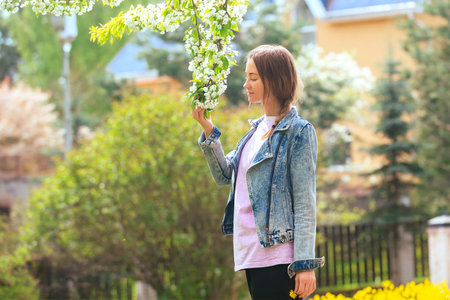 a slender beautiful blonde girl on the lawn near the house enjoys flowering trees and inhales their aromaの写真素材