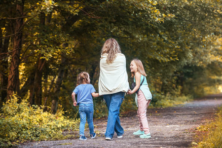 mom and her two little daughters hold hands and walk together along the path of the autumn park. family active vacation on warm autumn daysの写真素材