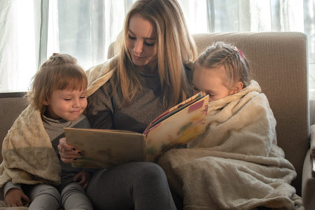 Mom at home on the sofa reads a book to her little daughters, they are wrapped in a blanket. Family...