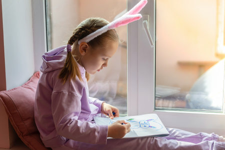 a schoolgirl girl with rabbit ears sits on the window and draws a rabbit in her album, preparing for Easterの写真素材