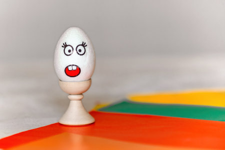 on colored paper, a stand with a white egg, on which there is a sticker with the emotion of surprise, fright, minimalism, copy space. communication on the Internet using emoticons with emotionsの写真素材