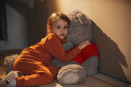 a little sad girl sits on the floor in an empty room, she hugged a huge teddy bear and looks thoughtfully somewhereの写真素材