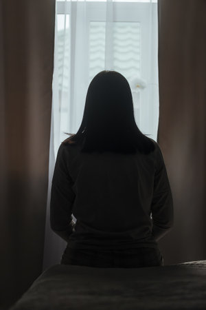 Silhouette of a young woman in pajamas against the background of a window at home on a bed with seasonal affective disorder or depression.. The concept of winter depression due to lack of sunlightの写真素材