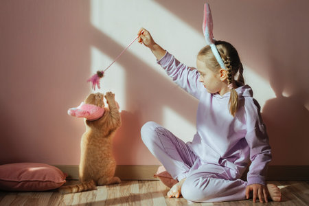 a schoolgirl girl on her head a headband with bunny ears plays with a red domestic cat in an Easter hat in the sunの写真素材