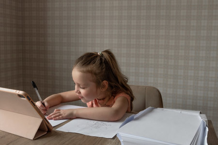 a little girl sits at a table in front of her a tablet and a folder of white paper, the child draws and copies from the tablet, online drawing lessonsの写真素材
