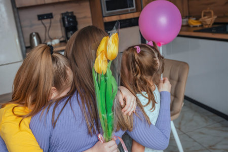 Daughters congratulate their mom on Mothers Day, a card with a heart, flowers and a balloon at home in the kitchen, they all hug. Children surprise their mother for the holiday.の写真素材