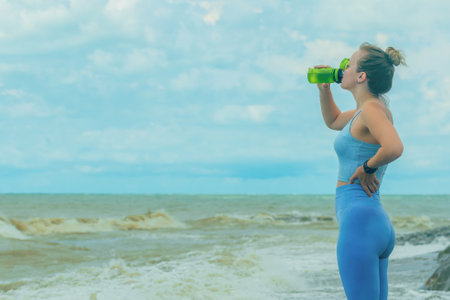 beautiful blonde sportswoman in a blue tracksuit during fitness on the seashore, drinks water from a shaker, copy spaceの写真素材