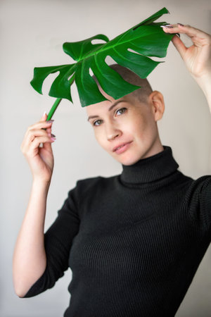 Portrait of an emotional young woman in a black turtleneck with a shaved head and a large green leaf of a flower behind which she hides. Outcome of chemotherapy in cancer treatmentの写真素材