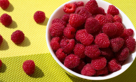 Raspberries in a white bowl on a green background. Ripe berries are scattered.