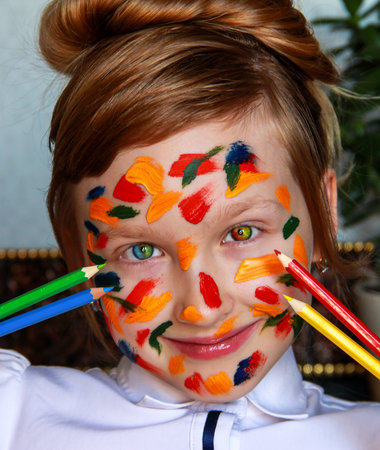 A girl stained with paint holds colored pencils.