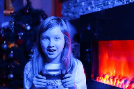 A girl plays a game console on the background of a Christmas tree and a fireplace. Gamer. New year's...