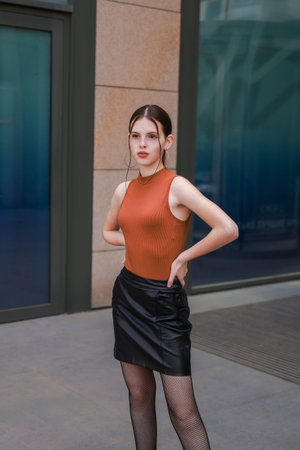 A girl with freckles in a leather skirt near a skyscraper.