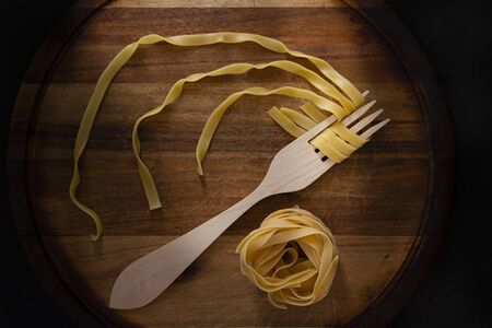 Round wooden board, on it a wooden fork and a nest of egg pasta, all in black backgroundの写真素材