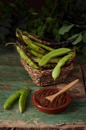 In the foreground, a ceramic bowl with carob flour. Around and at the bottom, pods and carob leaves. All on black background. Montuiri, Mallorca, Spainの写真素材