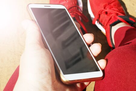 Hand with a smartphone on the background of feet and red sneakers, so closeの写真素材