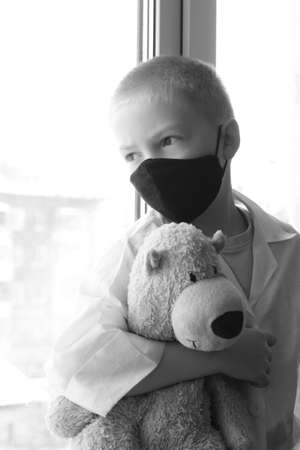 A little boy in a black mask and white bear toy at the window, self-isolation and quarantine, coronavirus. A portrait of a child.の写真素材