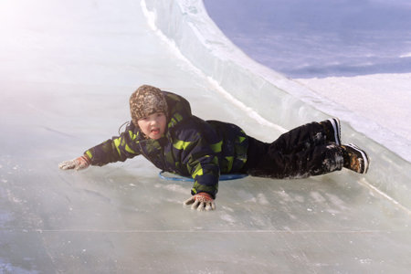 Little boy enjoy riding on ice slide in winter. Outdoor winter activities for family with kids.の写真素材