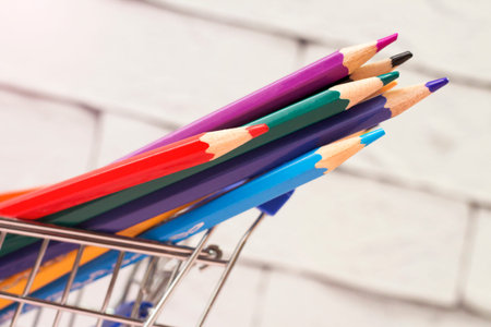 Colorful pencils in shopping cart on white brick wall background.