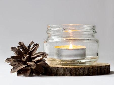A burning candle in a glass jar on a white background with a pine coneの写真素材