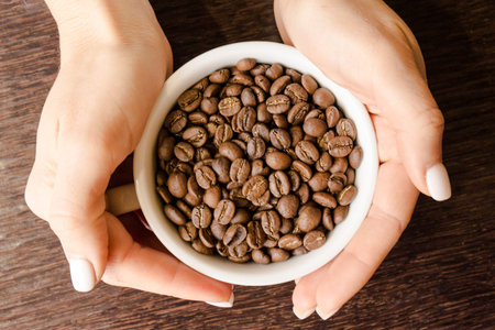 Fresh roasted coffee beans in a white Cup. Hands with a Cup of coffee on the background of a brown...