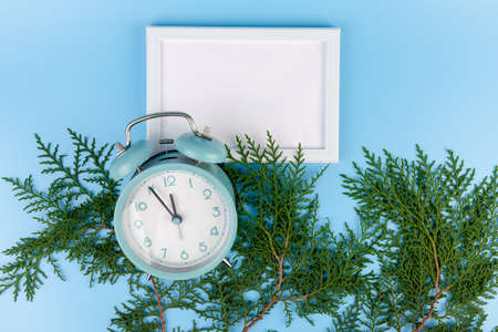 Blank white photo frame, blue alarm clock and green cypress branch on blue background. Christmas composition, copy space.の写真素材